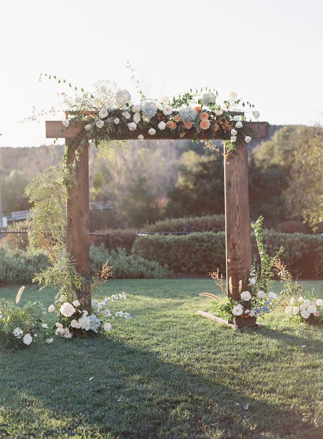 an outdoor ceremony setup with flowers and greenery on the grass in front of a wooden arch