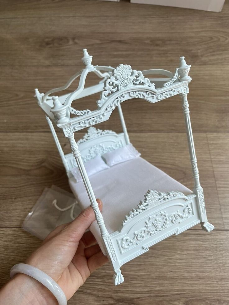 a hand holding a miniature white bed on top of a wooden floor