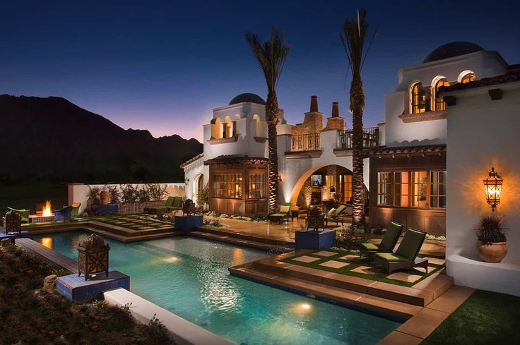 an outdoor swimming pool in front of a house with palm trees and lights on it