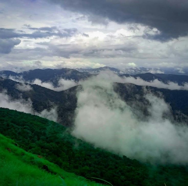 the clouds are rolling over the mountains in the valley below, and green grass is growing on the hillside