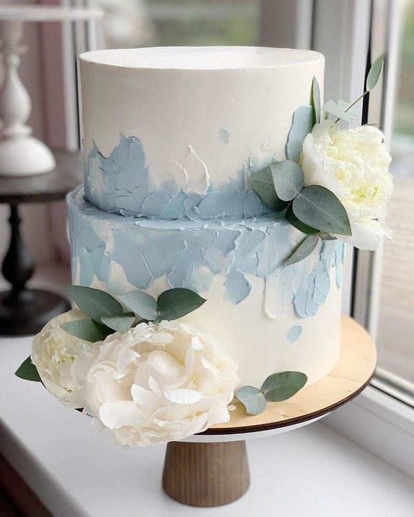 a white and blue wedding cake with flowers on the top is sitting in front of a window