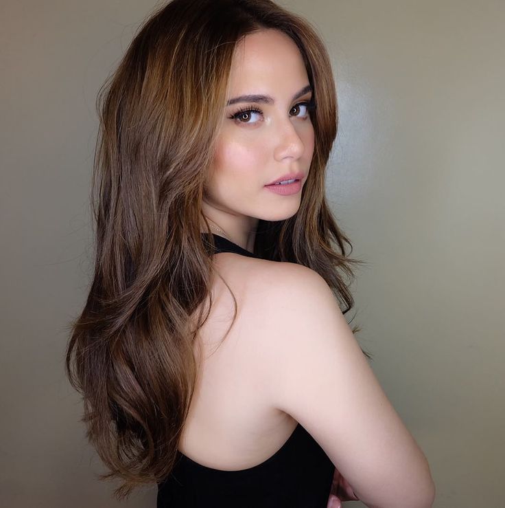 a woman with long brown hair wearing a black dress and looking at the camera while standing in front of a white wall