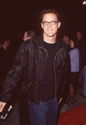 a man wearing glasses and a black jacket standing on a red carpet with people in the background