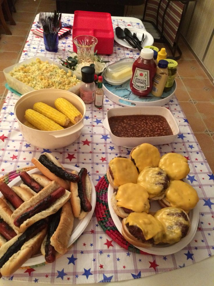 a table topped with hot dogs, corn and cheese covered hamburgers on buns