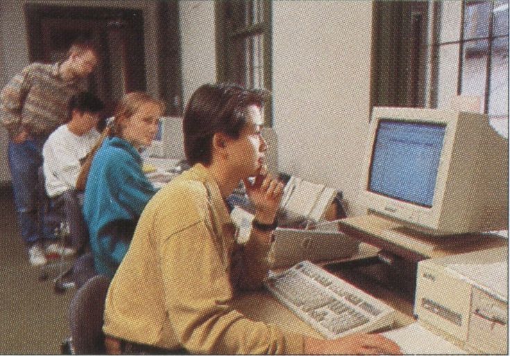 several children sitting in front of computers and talking on cell phones while one boy sits at the desk