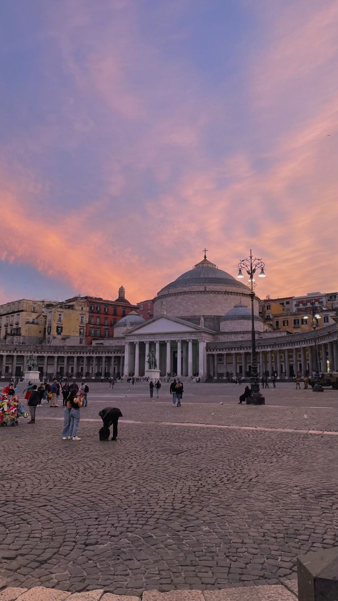 people are walking around in front of a building with columns and pillars on it at sunset