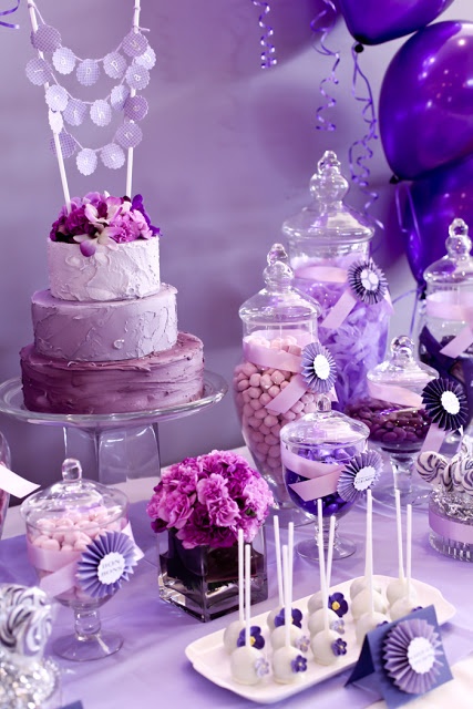 a table topped with lots of purple and white desserts next to balloons in the air