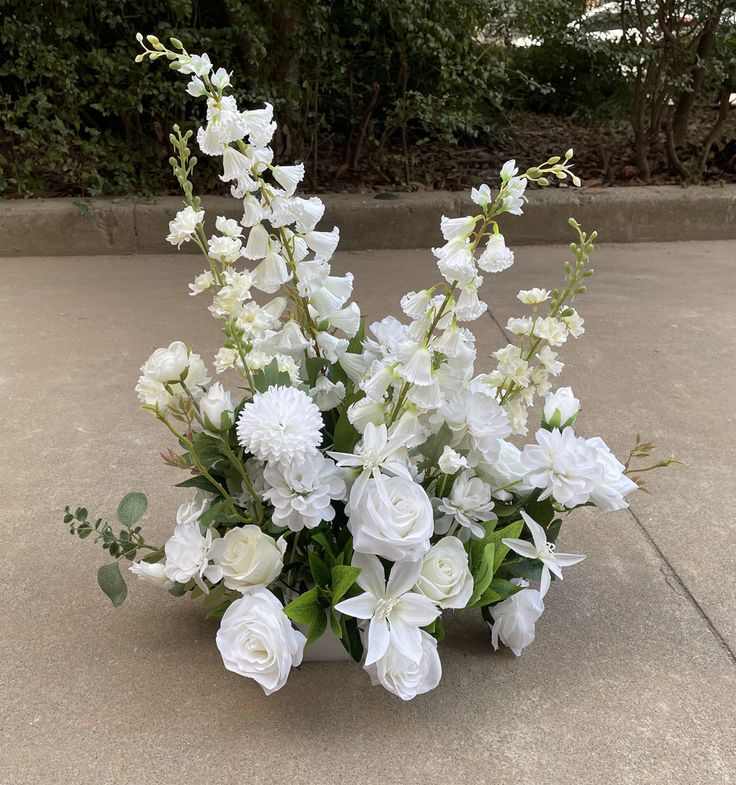 white flowers and greenery are arranged in a square vase on the concrete floor outside