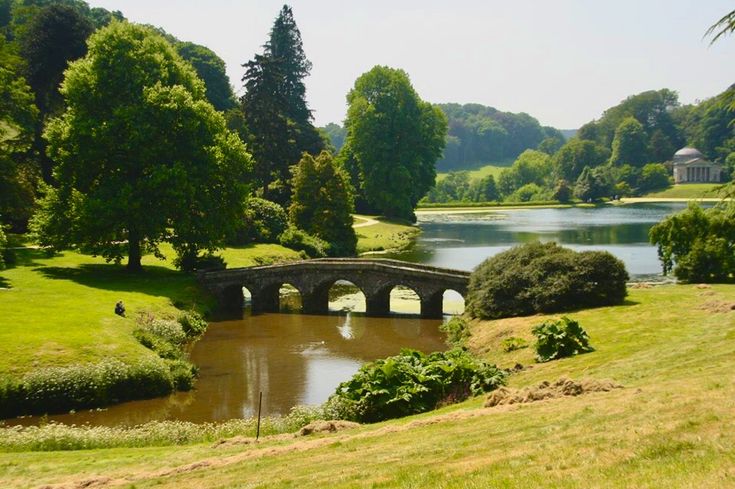 a stone bridge over a small pond in the middle of a lush green field with trees