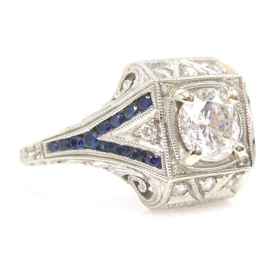 Rings like this are why we do what we do!This stunning diamond ring, made in our favorite Art Deco style, sports a 1.40ct E color diamond bead set at the center of a square setting. On each side of the diamond is a "Y" shaped channel, set with twelve natural blue sapphires. In addition to the sapphires, there are three round brilliant cut diamonds bead set in each beveled end and one round brilliant cut diamond in each shoulder.The piece is made of 14K white gold.DETAILS (CENTER STONE)__________ White Diamond Sapphire Ring, Gia Certified, White Sapphire Diamond Ring Gia Certified, White Sapphire Ring With Gia Certified Diamond, Art Deco White Sapphire Ring With Prong Setting, Art Deco White Diamond Ring With Prong Setting, White Art Deco Diamond Ring With Prong Setting, Engagement Ring With Sapphire Accents, Diamond Engagement Ring With Sapphire, Engagement Ring With Sapphire