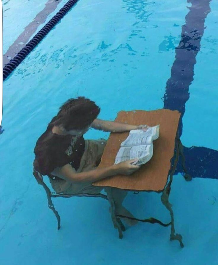 a woman sitting at a table in the middle of a swimming pool reading a paper