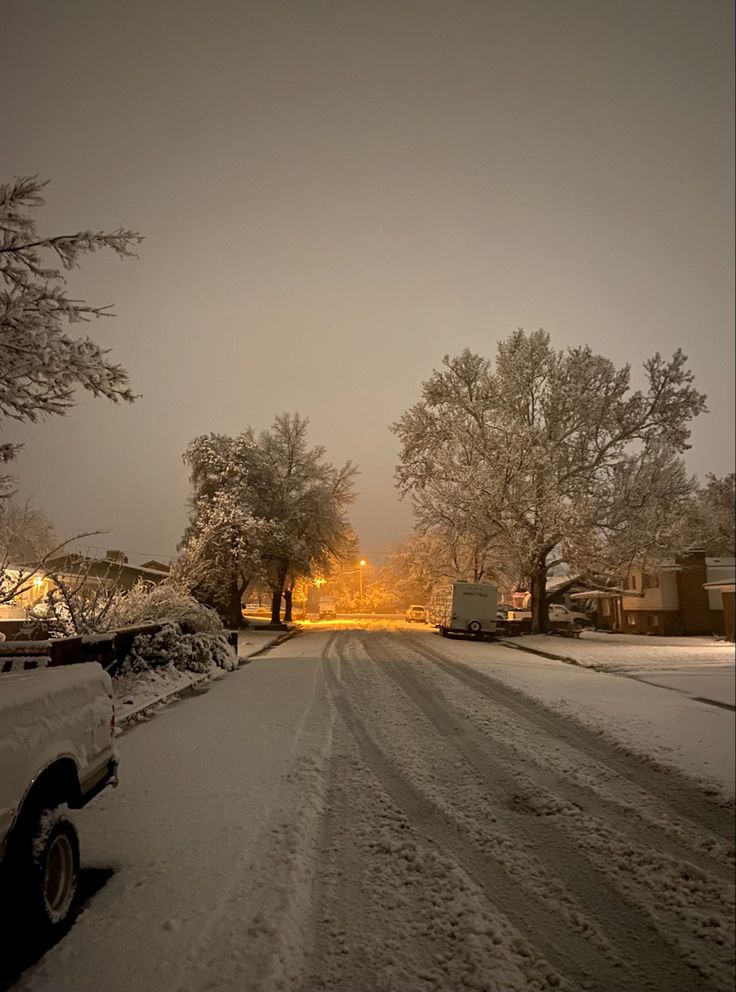 a snow covered street with cars parked on the side and houses in the background at night