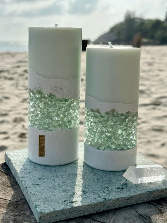 two white candles sitting on top of a rock near the ocean with sea glass in between them