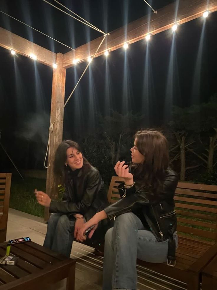 two women sitting on a wooden bench talking to each other at night with lights strung overhead