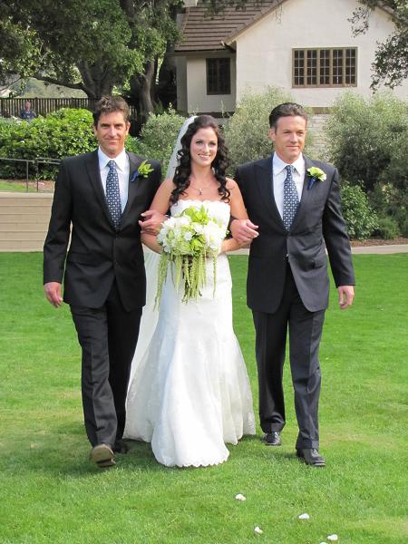 two men and a woman are walking in the grass with one man holding a bouquet