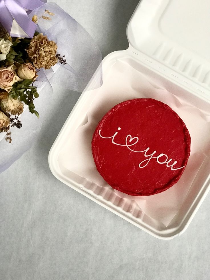 a red cake with the word i love you on it sitting in a plastic container next to flowers