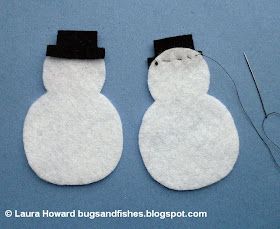 two felt snowmen sitting next to each other on a blue surface with sewing needles