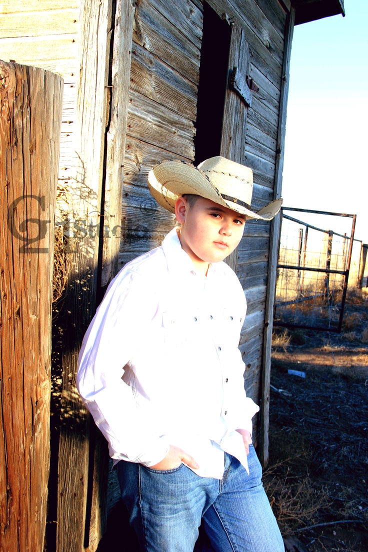 a young boy wearing a cowboy hat leaning against a wooden building with his hands on his hips