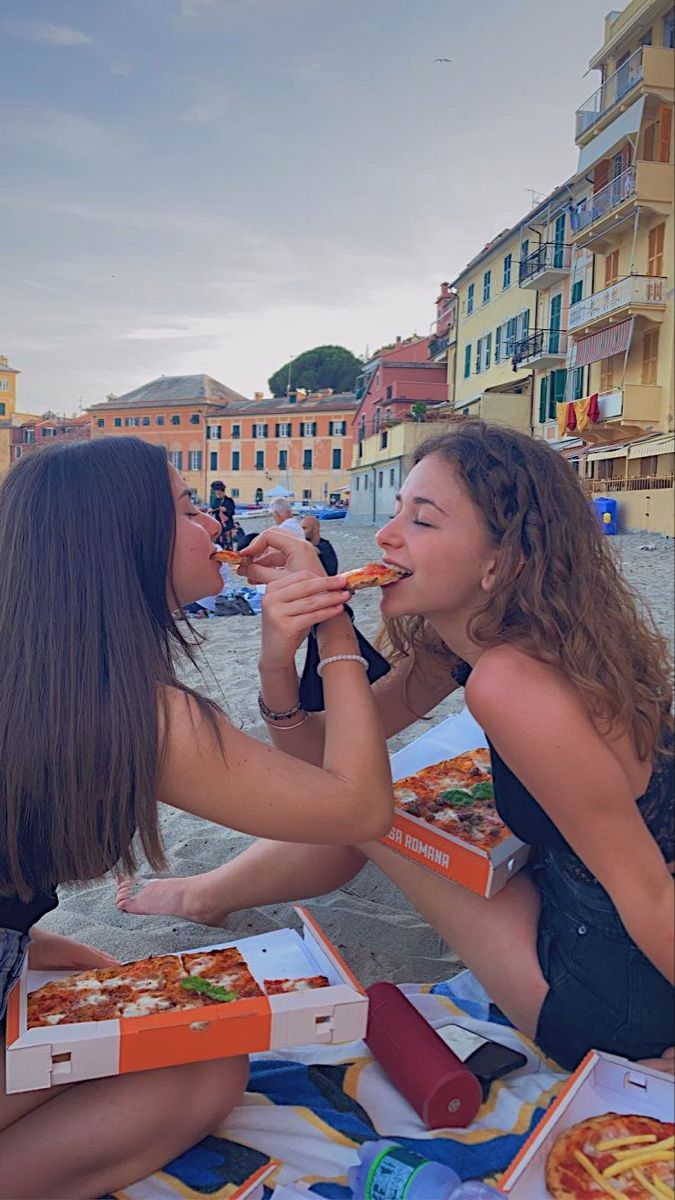 two women sitting on the ground eating pizza and drinking water from their mouths, with buildings in the background