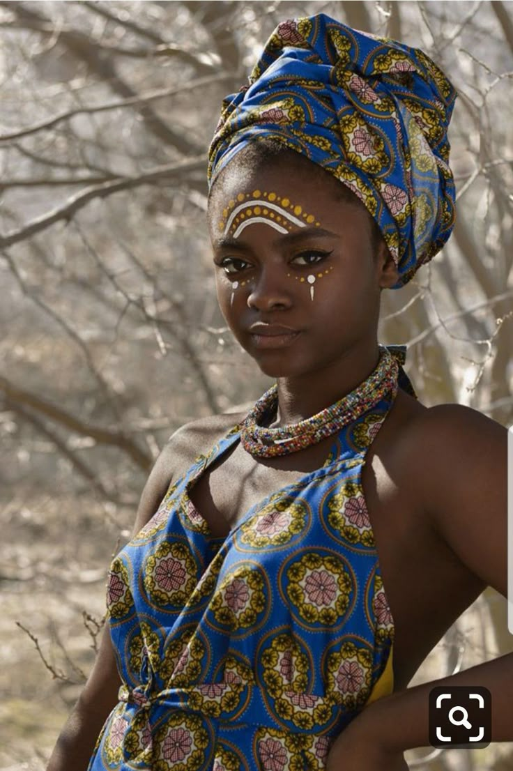 a woman in a blue and yellow dress standing next to some trees with her nose ring on