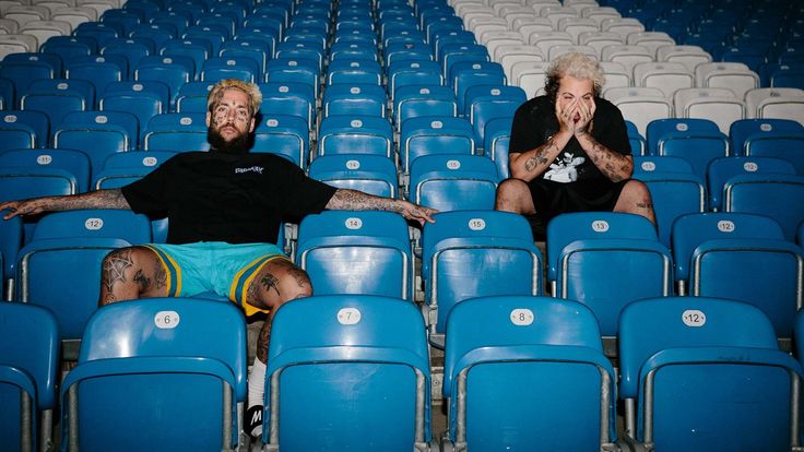 two men sitting on blue chairs in an empty stadium with their hands up to their mouths