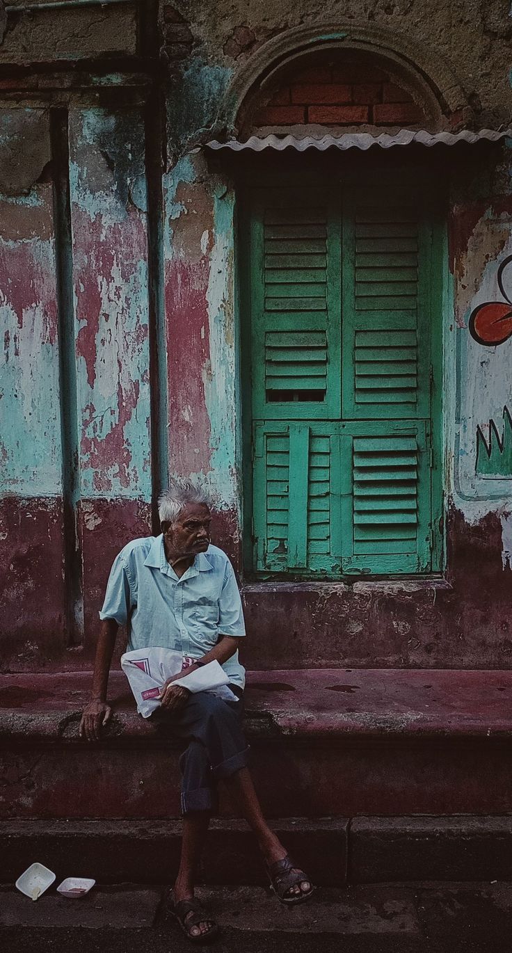 an old man sitting on the steps in front of a building with green shutters