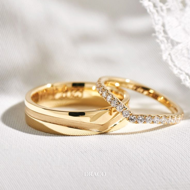 two gold wedding rings with diamonds on top of each other, sitting on a white cloth