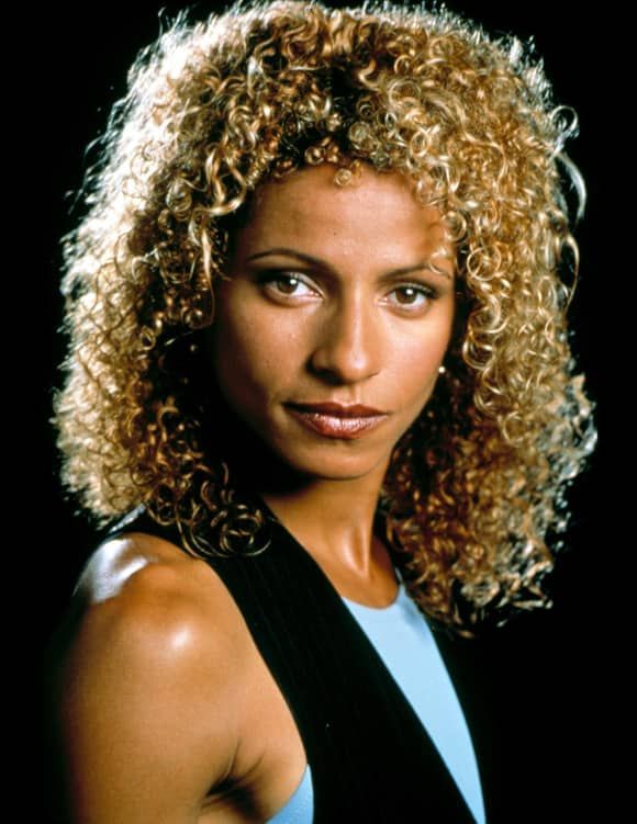 a woman with curly hair and blue shirt looking at the camera, in front of a black background