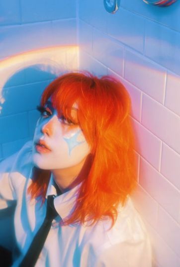 a woman with red hair and face paint sitting in front of a white tiled wall