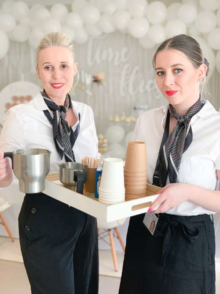 two women in aprons holding trays with food on them and balloons behind them