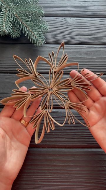 two hands are holding an ornament made out of wood and paper, with pine branches in the background