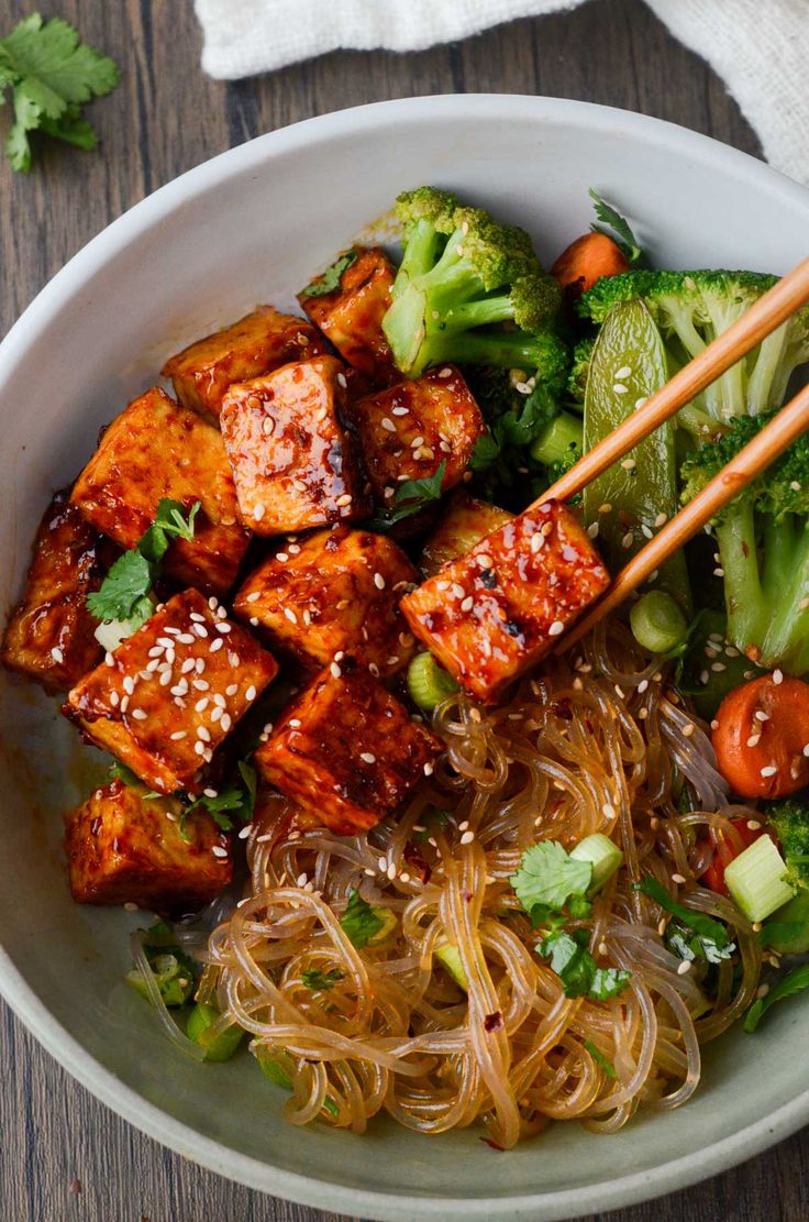 a bowl filled with noodles, broccoli and tofu on top of a wooden table