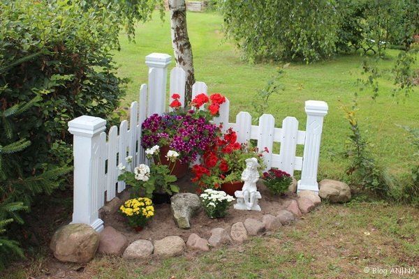 a white picket fence surrounded by flowers and rocks on the ground in front of a tree