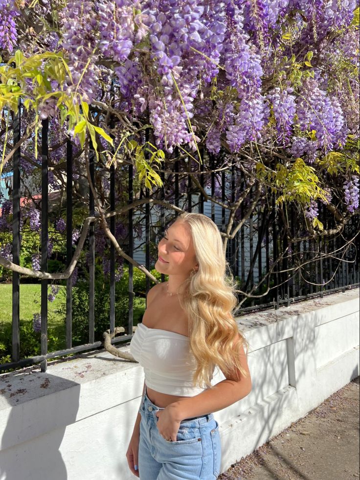 a woman standing next to a fence with purple flowers on the trees in front of her