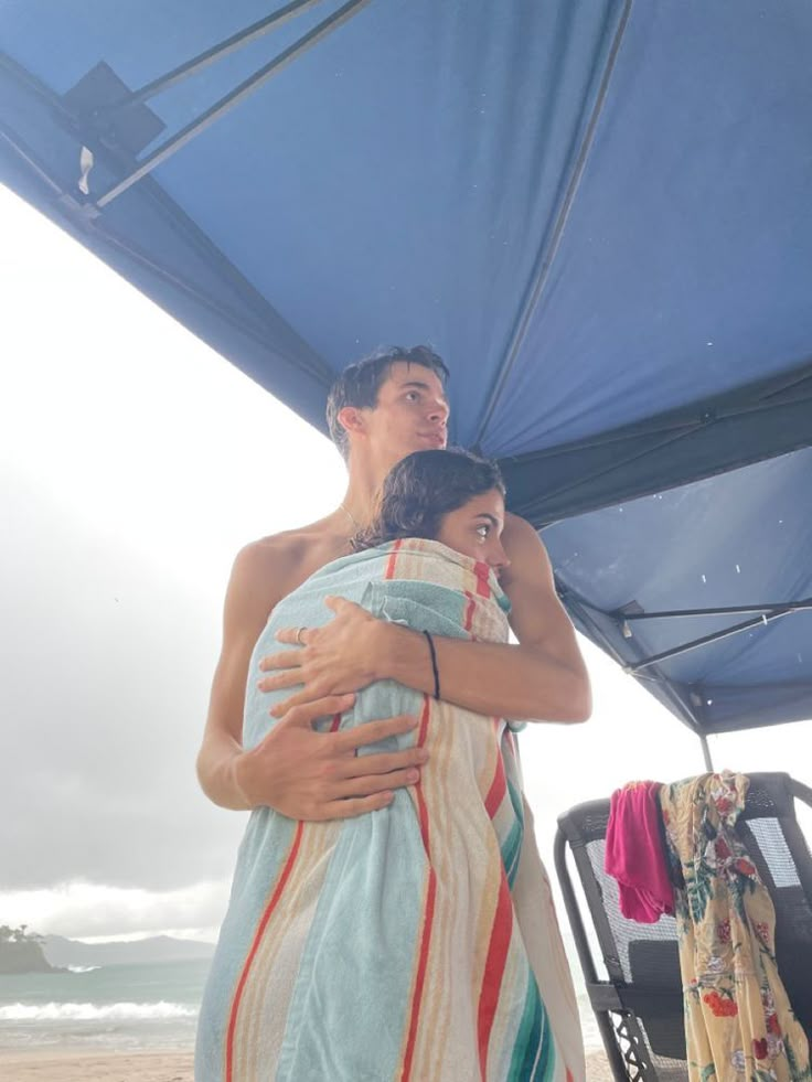 a man and woman wrapped up in towels under an umbrella on the beach, embracing