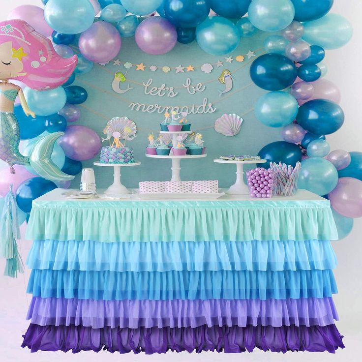 a table topped with lots of blue, purple and green balloons next to a cake