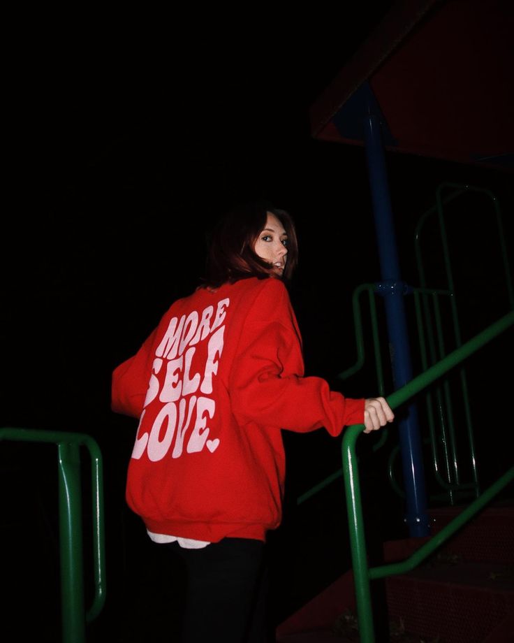 a woman wearing a red sweatshirt and black leggings stands on some stairs at night