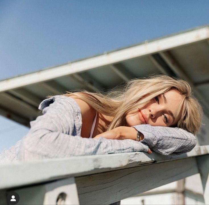 a beautiful blonde woman leaning on the edge of a wooden structure with her arms crossed