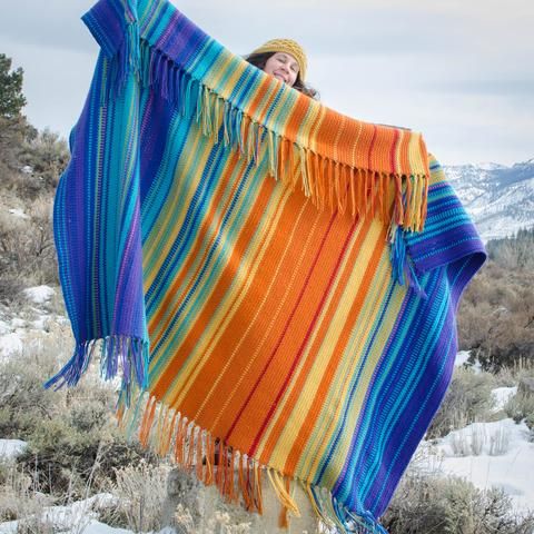 a woman wrapped in a colorful blanket on top of a snow covered hill with mountains in the background