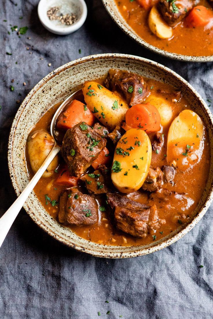 two bowls filled with stew and potatoes on top of a gray table next to spoons