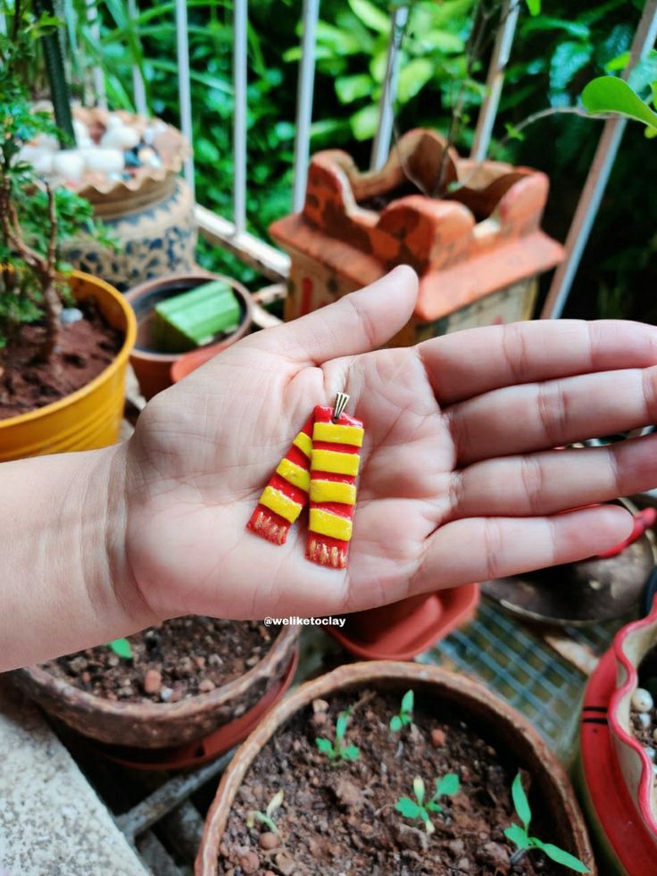 a person's hand holding two small yellow and red earrings in front of some potted plants