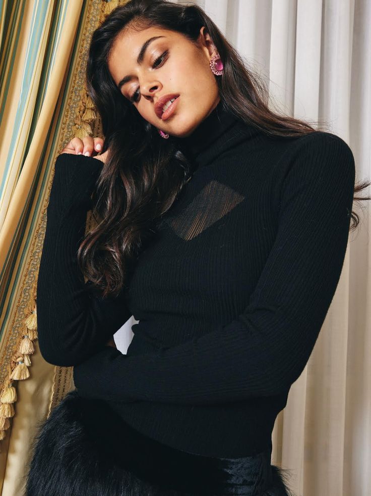 a woman posing in front of a curtain with her hand on her chin and wearing a black sweater