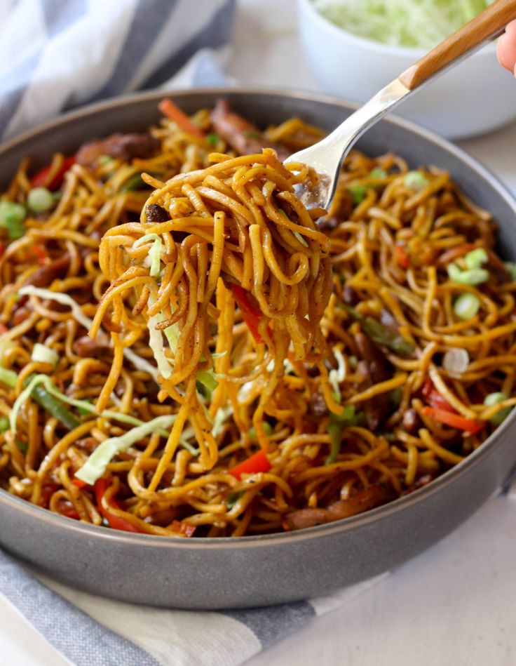 someone holding a spoon full of noodles with vegetables and sauce in the bowl behind them