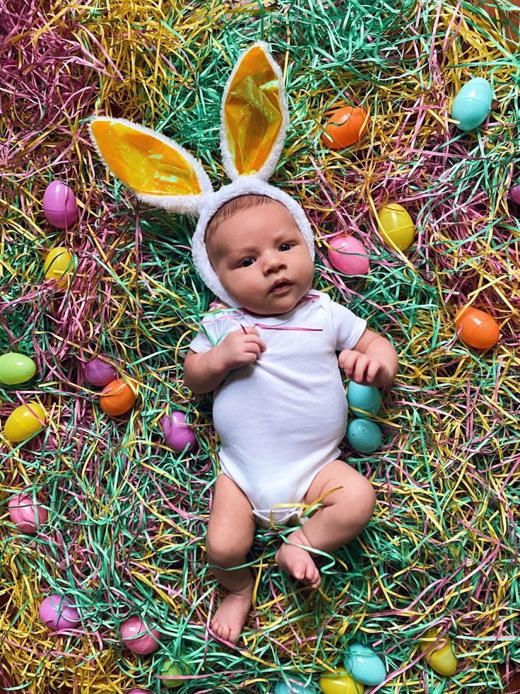 a baby wearing an easter bunny costume laying on some grass with colored eggs around it