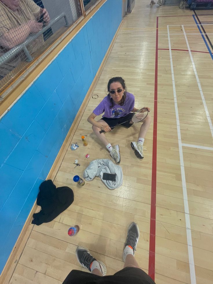 a woman sitting on the floor surrounded by shoes and other items in an indoor gym