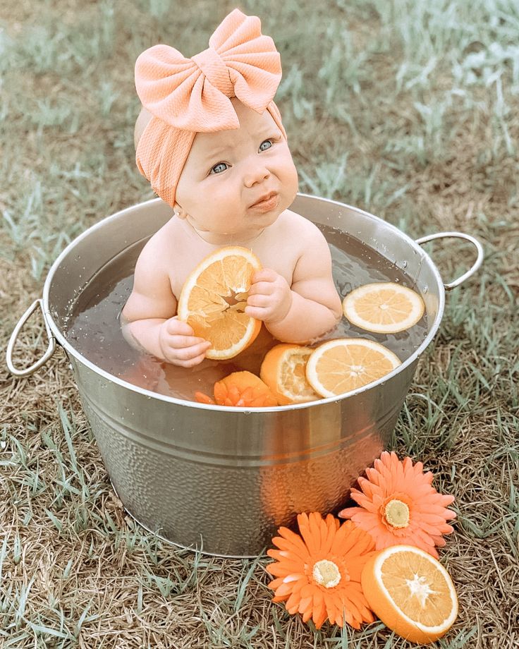 a baby sitting in a metal tub with oranges on the ground and flowers around it