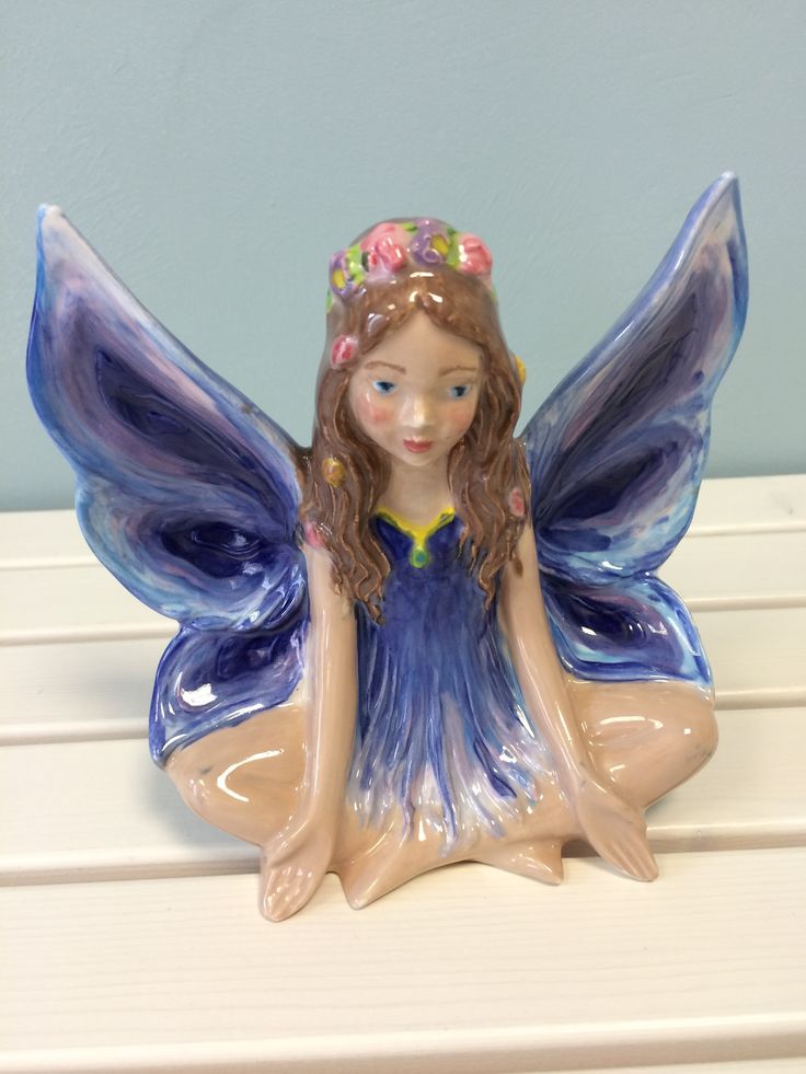 a ceramic figurine of a fairy sitting on a white surface with blue wings