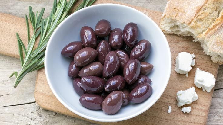 a bowl filled with olives next to bread on a cutting board