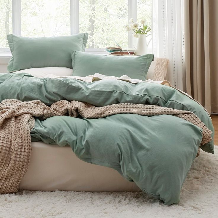 an unmade bed in front of a window with green sheets and blankets on it