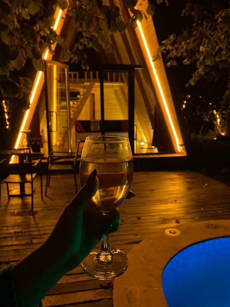 a person holding up a wine glass in front of a pool at night with lights on